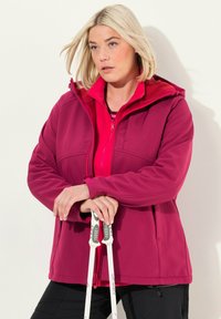 Fuksia pehmeäkuorinen takki, jossa on vetoketjullinen etuosa ja huppu, punaisen sisätakin päällä. Mallilla on valkoiset ja harmaat vaellussauvat kädessään.