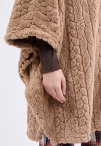 Chlupatý béžový pončo s texturovaným kabelovým pletením. Měkký plyšový materiál, široké rukávy a lem, který elegantně splývá.