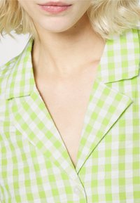 Camisa xadrez verde e branca, com gola e fecho de botão. O tecido parece leve, com uma ligeira textura e um design casual.
