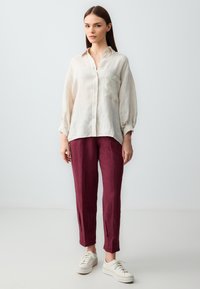 Camicia di lino color beige chiaro con tasca, abbinata a pantaloni di lino color bordeaux scuro. Sneakers bianche con suola testurizzata completano l'outfit.