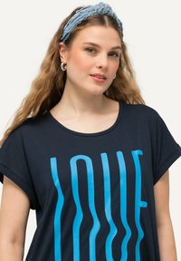 Camiseta de color azul marino de manga corta, con un gráfico ondulado de "AMOR" en azul brillante. Tejido suave con un corte relajado. Accesorio de diadema de mezclilla.