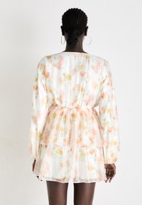 Vestido de tela ligera con estampado floral, con volantes, mangas largas y cintura elástica. Presenta colores pastel de flores rosas y amarillas sobre un fondo blanco.