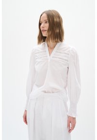 Weiße Bluse mit V-Ausschnitt, die dekorative Cut-Out-Details und geraffte Ärmel aufweist. Aus leichtem Stoff gefertigt, der eine weiche Textur schafft.