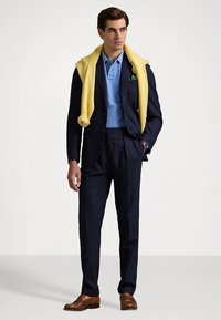 Ανδρικό μπλε ναυτικό κοστούμι με λεπτές ρίγες, ανοιχτό μπλε πόλο και κίτρινο πουλόβερ περασμένο πάνω από τους ώμους. Καφέ δερμάτινα παπούτσια.