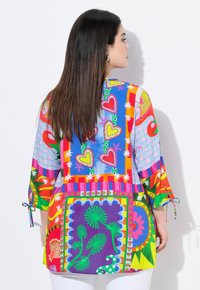 Tunique multicolore avec des motifs audacieux, comprenant des cœurs, des étoiles et des motifs floraux. Fabriquée en tissu léger, avec des manches 3/4 et des accents noués.