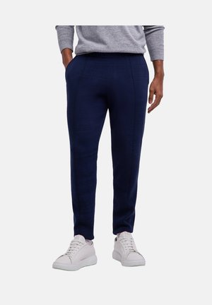Mann trägt marineblaue Slim-Fit-Hosen und weiße Sneakers, mit einer Hand in der Tasche und grauem Pullover sichtbar oberhalb der Taille.