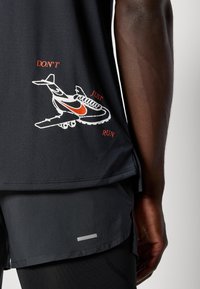 Fekete sportos tank-top, repülőgép és cipő grafikai mintával. Piros szöveg: "NE CSAK FUSS!" Kontrasztos varrásokkal és oldalhasítékokkal rendelkezik.