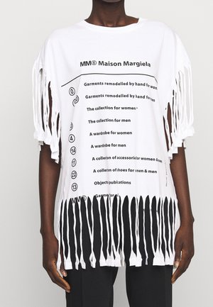T-shirt blanc oversize Maison Margiela avec texte imprimé en noir et détails de franges aux manches et à l'ourlet, porté avec un pantalon noir.
