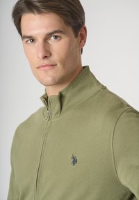 Felpa zip-up verde oliva con colletto alto, dotata di un piccolo logo blu navy sul petto. Texture liscia e design aderente.