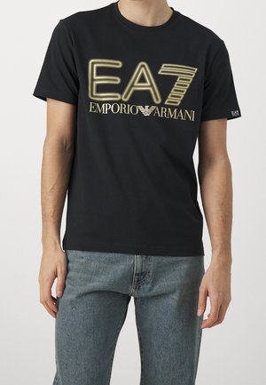 Man draagt een zwarte Emporio Armani EA7 T-shirt met gouden letters en lichtblauwe spijkerbroek, staand tegen een effen achtergrond.
