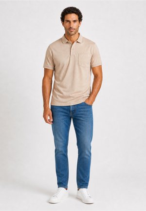 Mann steht und trägt ein beige kurzärmliges Poloshirt, blaue Jeans und weiße Sneakers mit einer Hand in der Tasche vor neutralem Hintergrund.