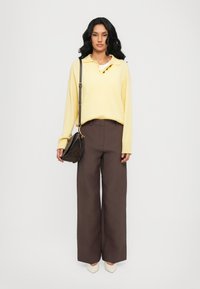 Polo a maniche lunghe giallo con bottoni, abbinato a pantaloni larghi marroni. Accessoriato con una piccola borsa scura e scarpe color crema.