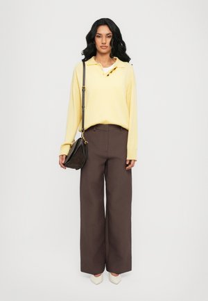 Yellow long-sleeve polo shirt with button placket, paired with brown wide-leg trousers. Accessorized with a small dark handbag and cream shoes.