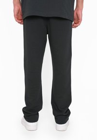 Black sweatpants with an elastic waistband, tapered legs, and soft texture, paired with white sneakers, viewed from the back.