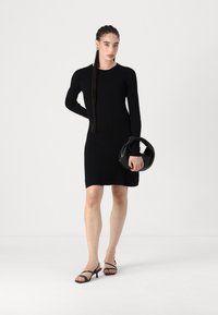 Μαύρο πλεκτό φόρεμα με μακριά μανίκια, στρογγυλή λαιμόκοψη και εφαρμοστή γραμμή. Συνδυάζεται με καμπυλωτή μαύρη τσάντα και μαύρες ψηλές γόβες με λουράκια.