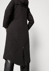 Manteau long noir avec capuche, comportant une fente latérale zippée et une texture de tissu lisse. Détails de coutures visibles au dos.