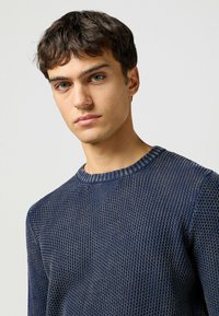 Marinblå stickad tröja med texturerat mönster, ribbad rund halsringning och subtila detaljer på ärmar och axlar.