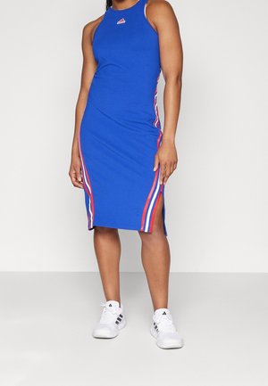 Femme portant une robe midi sans manches bleue avec des bandes latérales rouges, blanches et bleues et des baskets blanches Adidas, debout devant un fond uni.