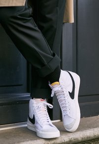 Weiße High-Top-Sneaker mit schwarzem Nike-Swoosh, grauem Zehenbereich und weißen Schnürsenkeln. Tragen Sie sie zu schwarzen, umgeschlagenen Hosen und gerippten schwarzen Socken.