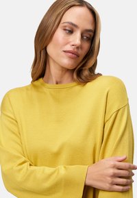 Pull jaune, coupe lâche, col rond, poignets côtelés, tissu texturé, manches longues, le mannequin inclina légèrement la tête.
