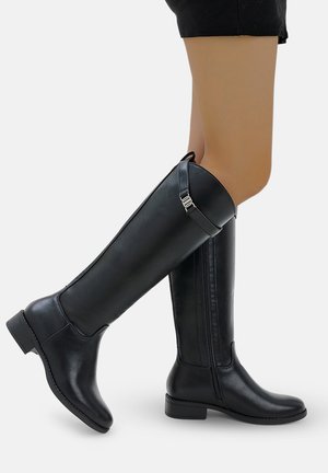 Piernas que llevan botas de cuero negras hasta la rodilla con cremalleras laterales y correas de hebilla, mostradas contra un fondo claro y liso.