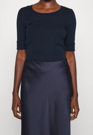 Femme portant un haut noir à manches courtes avec des boutons dorés aux épaules et une jupe en satin foncé sur fond blanc uni.