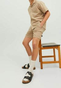 Homme portant un short beige et une chemise à manches courtes, chaussettes blanches, sandales noires, assis sur une chaise en bois devant un fond uni.