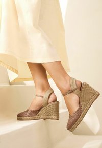 Brown woven wedge sandals with a textured upper, rope sole, and adjustable ankle strap. The heel is approximately 7 cm high.