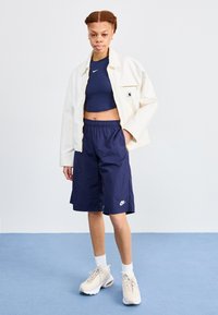 Bijela jakna s jednim džepom, plavi crop top i mornarsko plave šorts hlače s Nike logom. Bež tenisice s teksturiranim đonovima i usklađene čarape.
