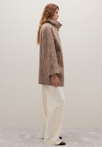 Brauner karierten Mantel mit hohem Kragen, strukturierter Stoff und überschnittenen Schultern, kombiniert mit weißen, weit geschnittenen Hosen und schwarzem Schuhwerk.