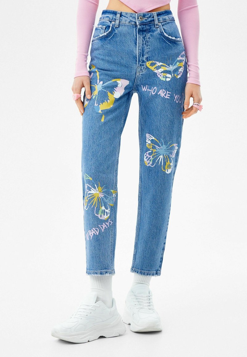 Ljusblå denimjeans med färgglada fjärilstryck, med texten "VEM ÄR DU?" och "INGA DÅLIGA DAGAR." Rakt skuren med standardfickor.