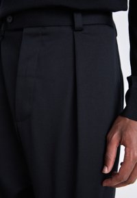 Gros plan sur un pantalon noir sur mesure avec une passants de ceinture et la main droite détendue d’une personne à côté de la cuisse.