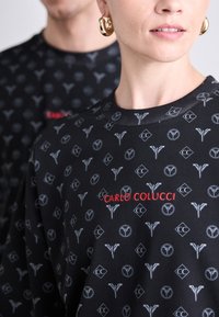 Sort sweatshirt med et gentaget gråt mønster og "CARLO COLUCCI" i rødt broderi. Rund hals med tekstureret detaljering.