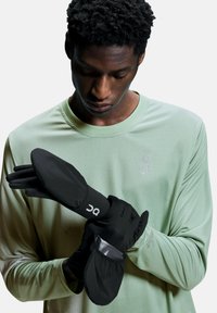 Gants de course noirs avec des poignets rembourrés et un accent transparent. Portés par une personne dans une chemise à manches longues texturée vert clair.