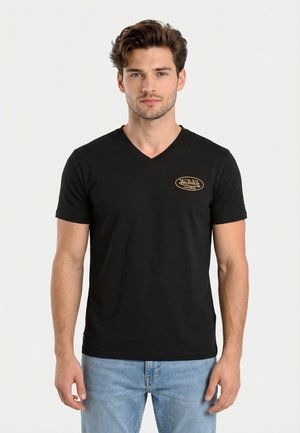 Hombre con cabello corto y castaño que lleva una camiseta negra de cuello en V con un pequeño logo amarillo de "Von Dutch California", combinado con vaqueros azul claro.