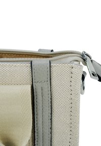 Close-up van de hoek van een beige canvas tas met een grijze leren schouderband en een gedeeltelijk open metalen rits op een witte achtergrond.