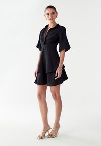 Vestido negro con mangas cortas y un cuerpo ajustado, falda en capas y tela texturizada. Combinado con sandalias blancas de tacón.
