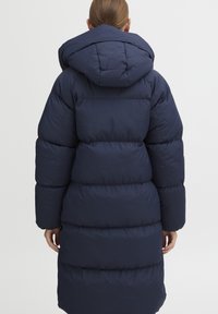 Marineblå puffjakke med hette, med quiltede seksjoner og lange ermer. Materialet ser glatt ut og isolert for varme.