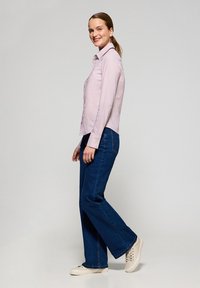 Camicia button-up viola chiaro con colletto, indossata con jeans flare blu scuro e sneaker beige, mostrata di profilo su uno sfondo neutro.