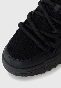 Black sneakers with a textured black fabric upper, featuring thick black laces and a smooth black leather toe cap and sole.