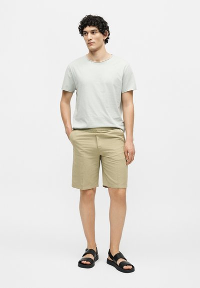 Νέος άνδρας στέκεται με τα χέρια στις τσέπες, φοράει ανοιχτό γκρι μπλουζάκι, μπεζ σορτς και μαύρα σανδάλια μπροστά σε λευκό φόντο.