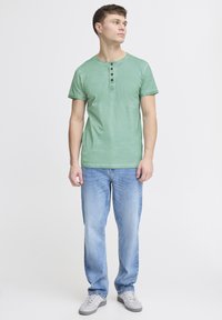 Grønn kortermet henley-skjorte med tre brune knapper, paret med lyseblå jeans og grå treningssko. Enkelt, uformelt design.