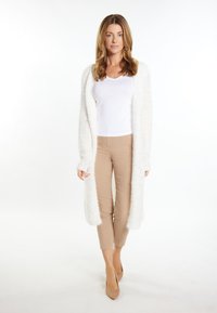 Lunga, bianca e morbida cardigan indossata sopra una maglietta bianca e pantaloni beige aderenti. L'outfit è completato da tacchi beige chiari.