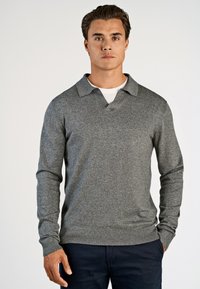 Mand iført en grå langærmet striktrøje over en hvid skjorte, parret med mørke bukser, stående foran en ensartet lys baggrund.