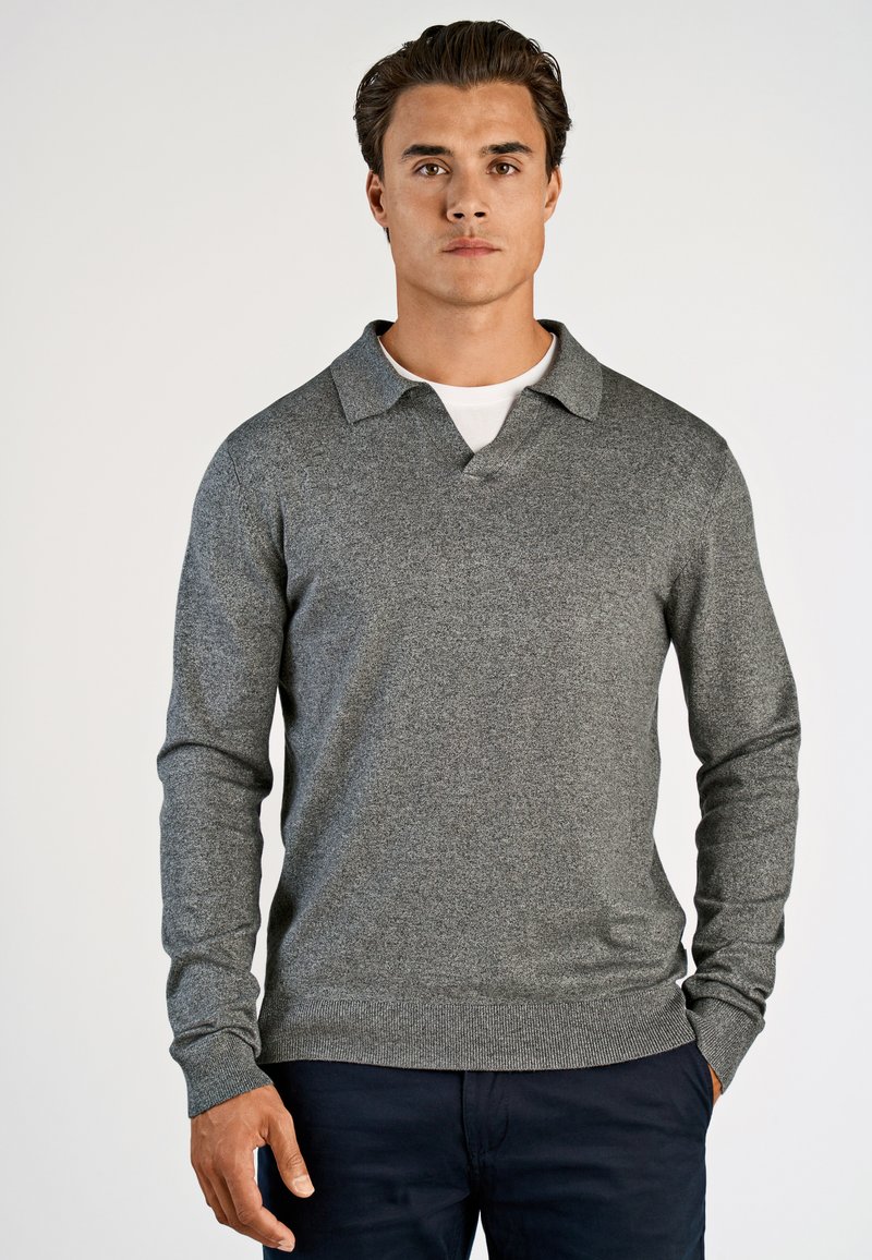 Mand iført en grå langærmet striktrøje over en hvid skjorte, parret med mørke bukser, stående foran en ensartet lys baggrund.