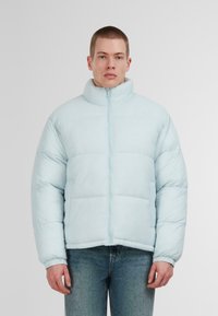 Šviesiai mėlyna pūkinė striukė su aukštu apykakle, pilnu užtrauktuku ir siunčių raštu. Yra elastiniai rankogaliai ir siauras siluetas. Pagaminta iš glotnaus, blizgaus medžiagos.