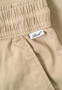 Beige elastic waistband pants with visible stitching and a small white fabric tag displaying "Reell" in black script font.