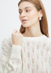 Weißer Strickpullover mit wellenförmigem Muster, gerippten Bündchen und weitem Ausschnitt; Model trägt einen silbernen Tropfohrring.