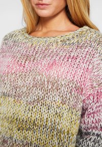 Close-up van een vrouw die een dikke, multiculaire gebreide trui draagt in roze, geel, wit en grijstinten.