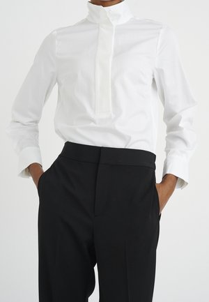 Hvid skjorte med lange ærmer, høj krave og knaplukning. Kombineret med sorte bukser med høj talje, der har en elegant og skræddersyet pasform.